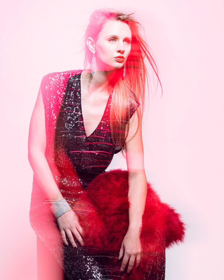 Fashion photography using colored gel and long exposure of young woman in stylish sequined jumpsuit sitting in fashion pose.